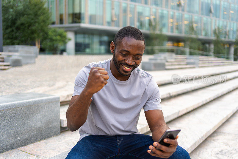 快乐的非裔美国人在城市街道上拿着智能手机欢欣鼓舞。人家伙看手机阅读伟大的新闻获得好结果赢得网上竞标感到惊讶，获胜的手势