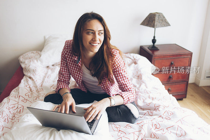 微笑的年轻女子坐在床上使用笔记本电脑