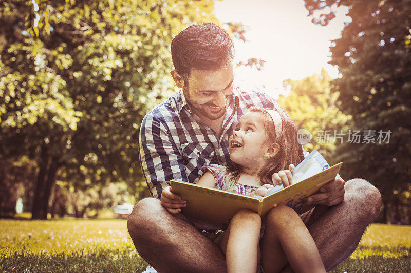 单身父亲和小女儿坐在草地上读故事书。小女孩坐在爸爸腿上。