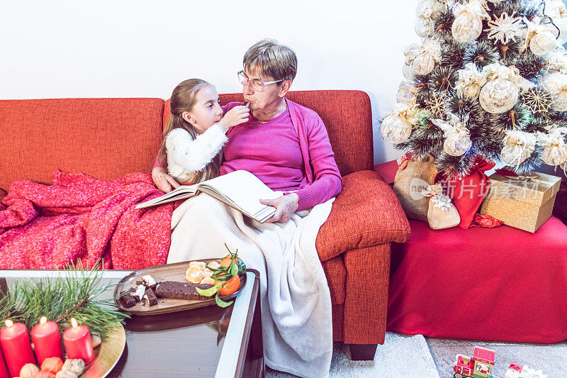 祖母和孙女在沙发上享用传统的圣诞牛轧糖(意大利Torrone)和橘子
