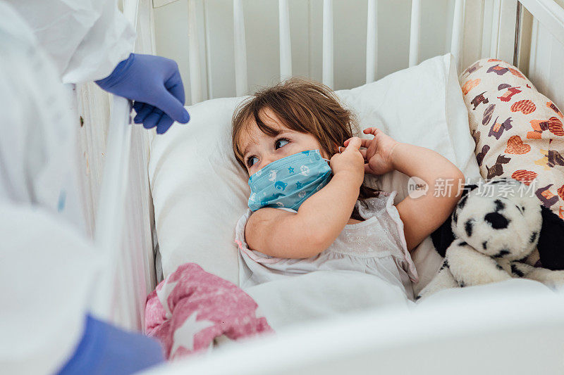 佩戴个人防护装备的医生在家中婴儿床上照料一位戴着医用口罩的金发小女孩