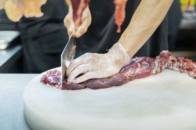 在一家牛肉火锅餐厅的开放式厨房里，厨师在挂着的切碎的生肉后面用刀切牛肉