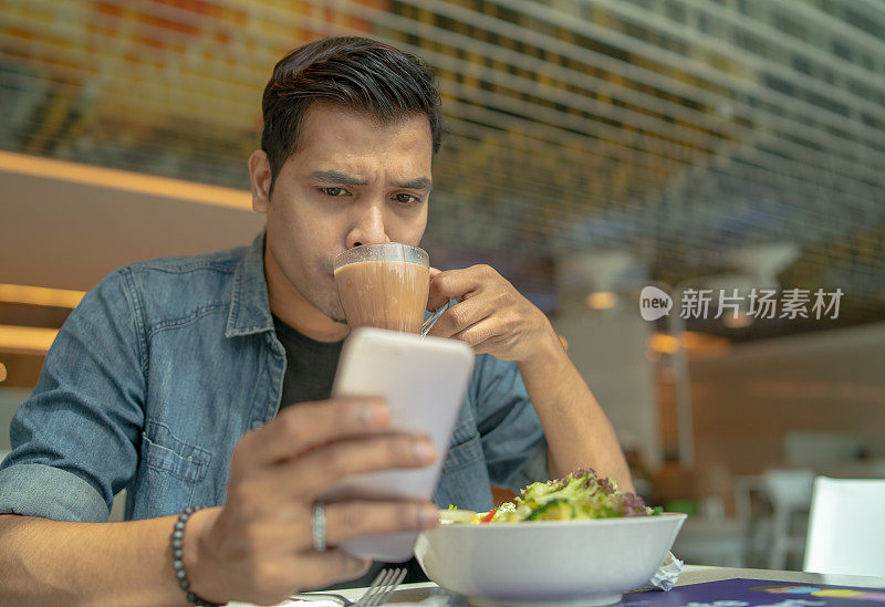 严肃的亚洲马来青年喝咖啡和使用手机