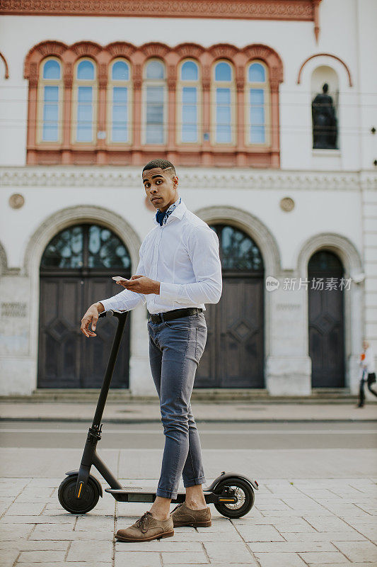 年轻的非洲裔美国人骑着电动车站在街上使用手机