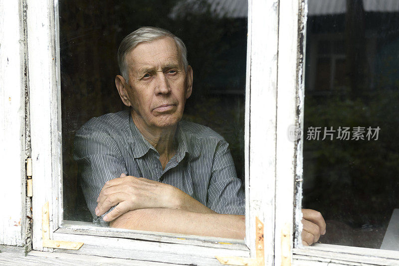 透过玻璃窗的肖像。一位悲伤的80岁老人坐在一座老房子里。