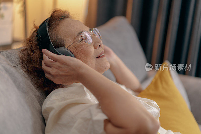 坦率的亚洲老年妇女坐在沙发上，戴着无线耳机听在线音乐从手机在家里放松。在客厅听音乐或播客。