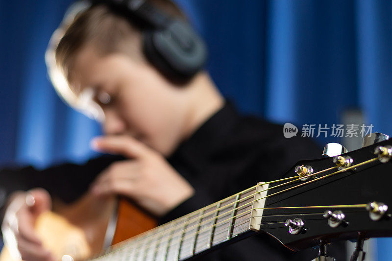 一个13岁的少年穿着黑色的衬衫，戴着耳机，在音乐工作室里用吉他在蓝色的背景上录制旋律。有选择性的重点。