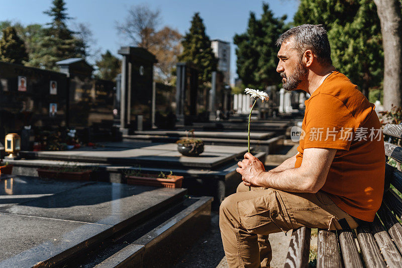 成熟悲伤的男人坐在墓地的墓碑旁，手里拿着一朵花，哀悼死者