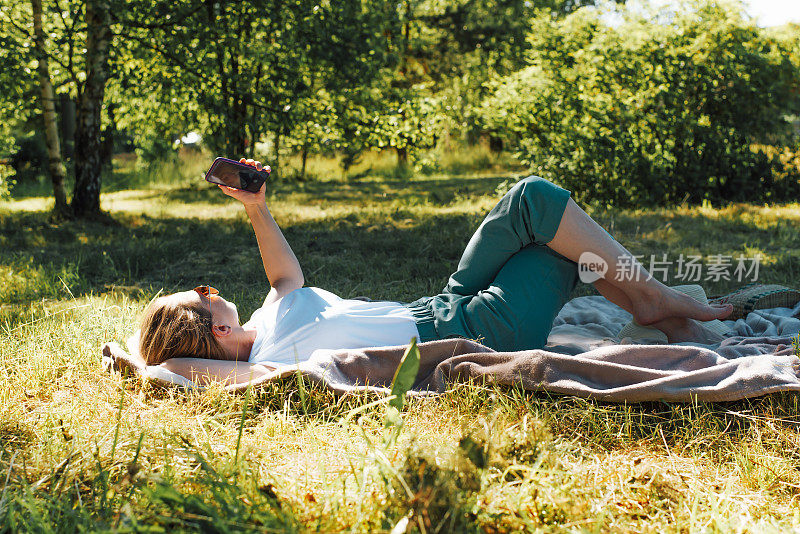 户外野餐，年轻女子躺在公园草坪上的毯子上，用智能手机拍自己的照片，侧视图。暑假女孩在大自然中休息