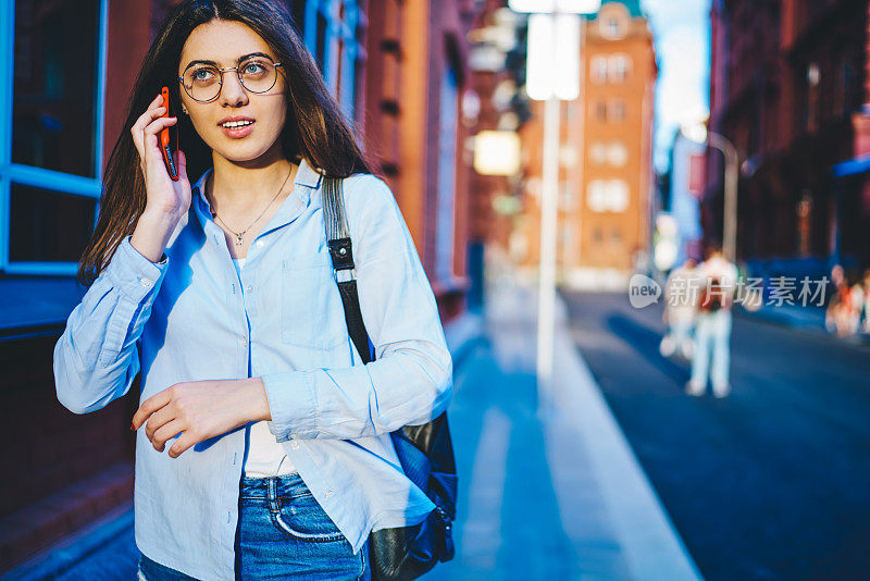 戴眼镜的年轻女子走在城市街道上打手机漫游，积极随意的女性在户外散步时聊天，满足于连接