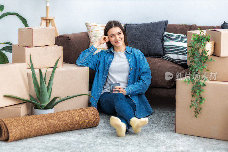 搬新家，租房子。快乐的年轻白人妇女坐在地板上与一杯咖啡和纸板箱在她的新家，微笑
