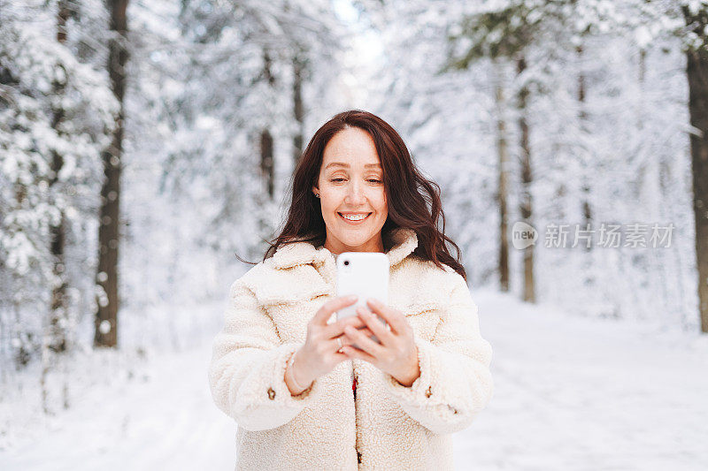 在白雪覆盖的冬季森林背景下，身穿冬装的黑发微笑女子正在使用手机