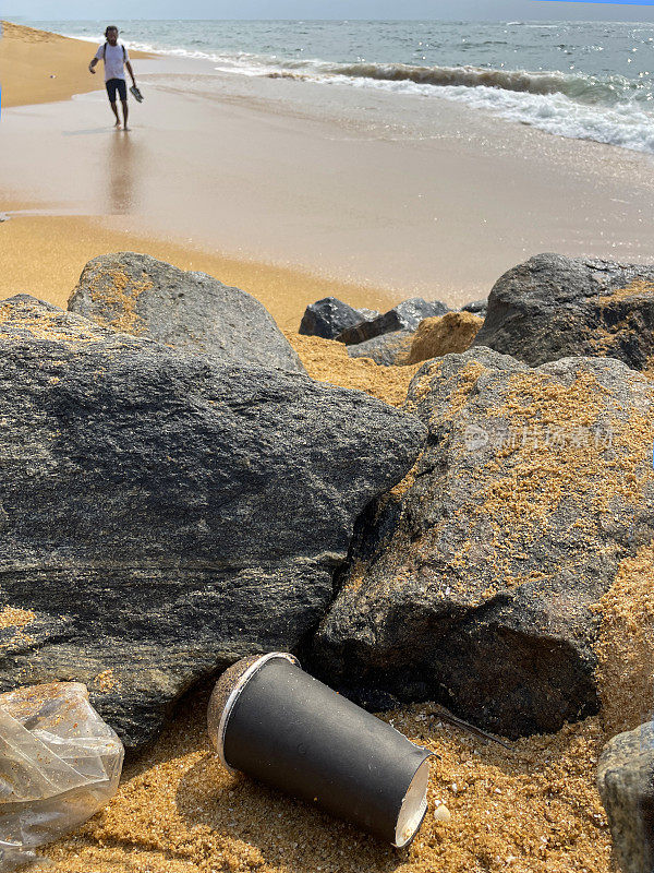 图像的空，纸板一次性咖啡杯与透明塑料圆顶盖冲在岩石池在退潮，海洋垃圾和污染的海岸，垃圾的沙子，肮脏的海滩，偶然的人，破碎的海浪背景，重点在前景