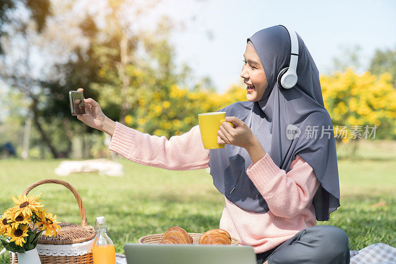 亚洲年轻的穆斯林妇女戴着希贾布头巾，用智能手机在假日周末公园野餐时快乐地微笑着拍照