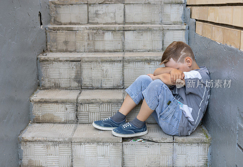 一个抑郁的孩子坐在楼梯上，双手抱着膝盖。夏天，一个孤独的男孩悲伤地把脸埋在膝盖里。一个没有朋友的孤独孩子的概念。