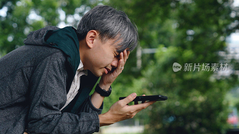 一位沮丧的亚洲男子坐在公园里接电话，脸上带着沮丧和悲伤的表情。