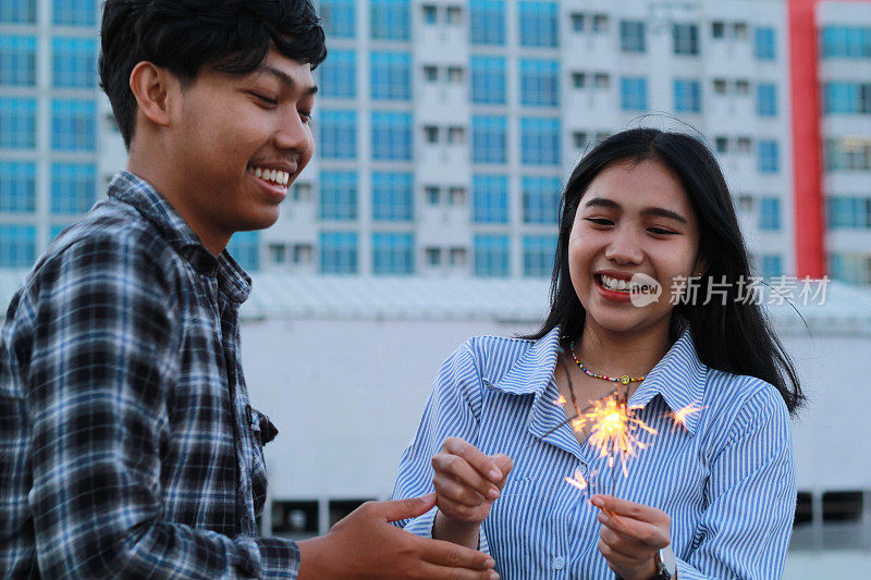 俏皮的亚洲年轻夫妇带着笑脸燃烧着烟花庆祝新年，在城市建筑背景下穿着法兰绒和条纹衬衫休闲