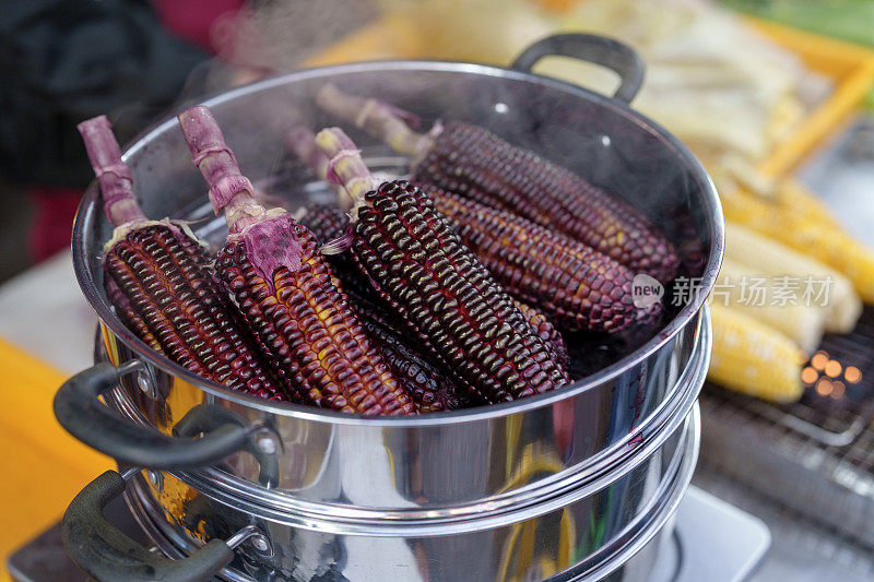 一个特写镜头，在一个亚洲夜市上，紫色的玉米被放在金属蒸笼上准备出售。