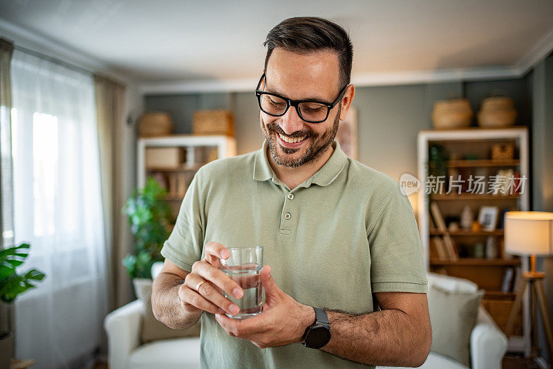 一个看起来年轻又成熟的男人站在客厅里，用杯子喝着新鲜干净的水，关心液体的摄入量