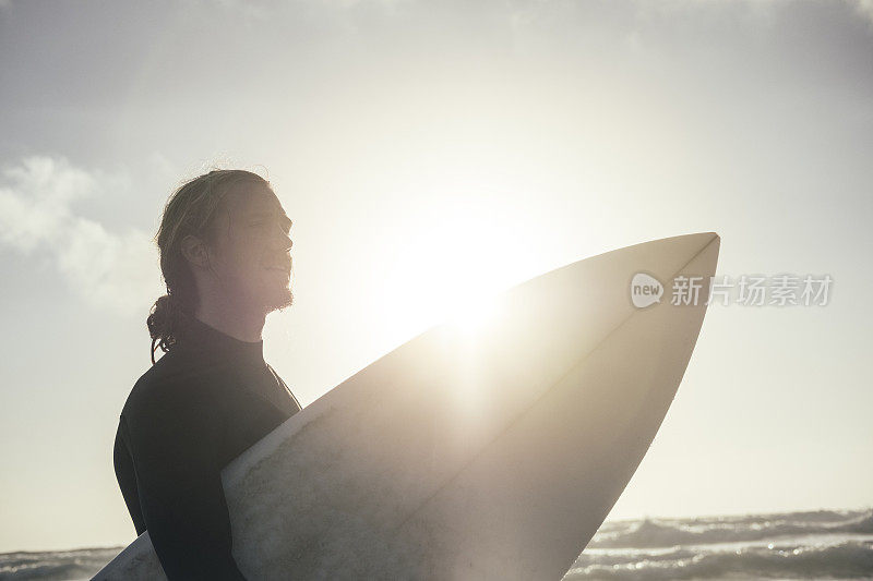 背光的冲浪者拿着冲浪板，带着夕阳光晕