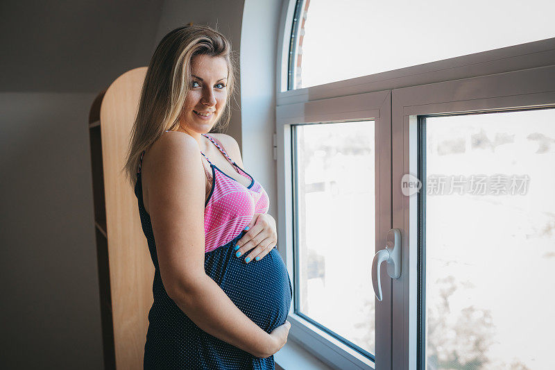 年轻快乐的怀孕母亲摆姿势旁边的窗户在家里和触摸她的肚子