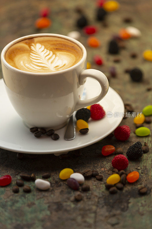 热咖啡拿铁杯与罗塞塔拿铁艺术牛奶泡沫，咖啡豆和彩色果冻糖果在质朴的木头桌子背景