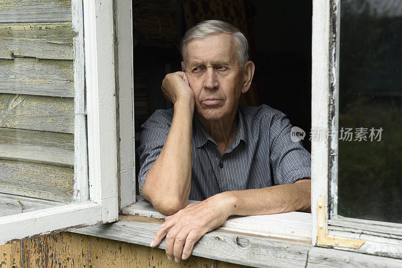 一位忧伤的80岁老人的肖像，坐在一座旧木屋的窗前。