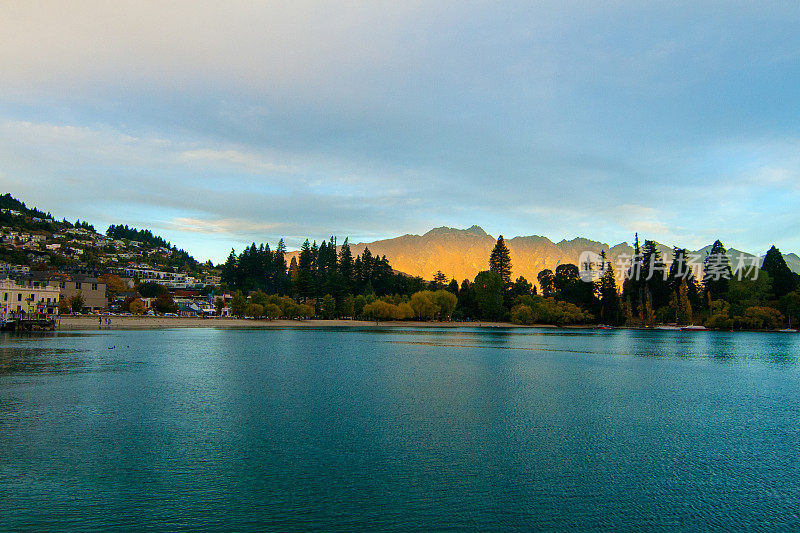 新西兰著名的度假小镇奥塔哥的皇后镇和标志性的山脉，蓝色的瓦卡蒂普湖之上的非凡山脉，浪漫的晚秋日落风景与金色的阳光