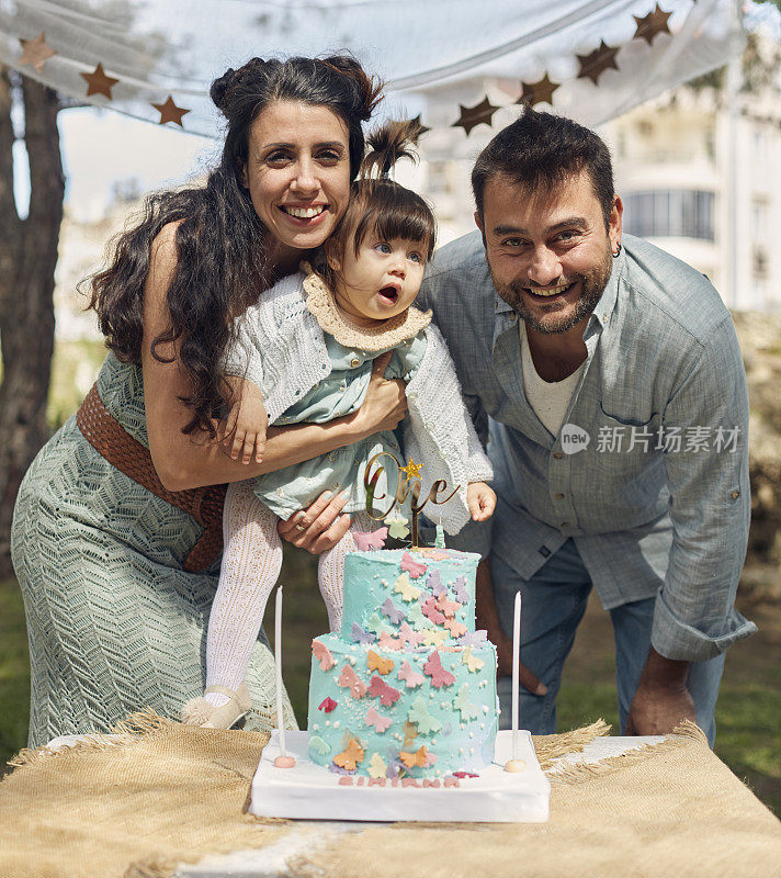 在花园里为小女儿庆祝生日，爸爸妈妈在生日蛋糕后面开心地摆姿势。