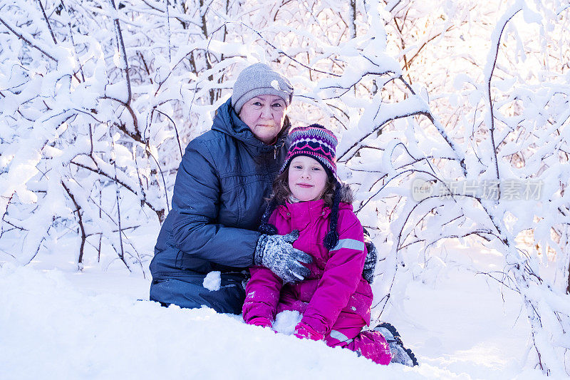 肖像。一位77岁的老妇人和一个7岁的女孩微笑着看着镜头，背景是白雪覆盖的树枝和阳光。