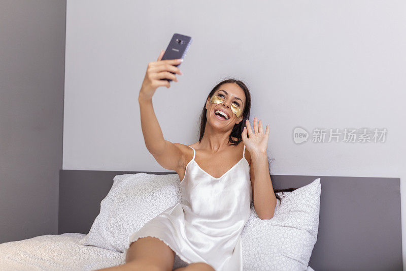 年轻快乐的女人带着金色化妆品面膜，眼罩用手机进行视频通话，手势亲吻朋友。漂亮的女孩躺在床上用智能手机进行facetime视频通话。