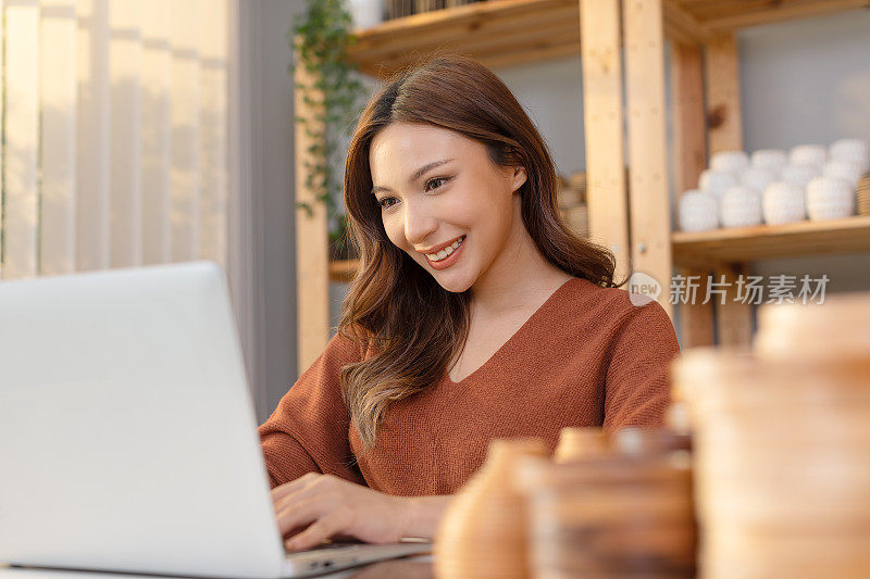 快乐的亚洲年轻企业主花瓶店工作和网上销售电脑笔记本电脑微笑。自信的女企业家在家里被美丽的花瓶包围。积极情绪。