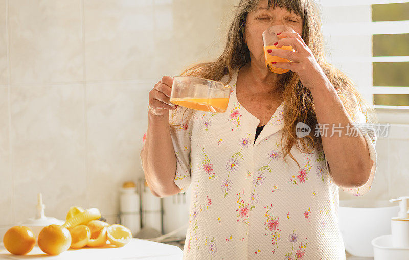 白种成熟的老年妇女喝一杯带果肉的鲜榨天然橙汁。早餐概念和早晨醒来。非常明亮明亮的家庭厨房。