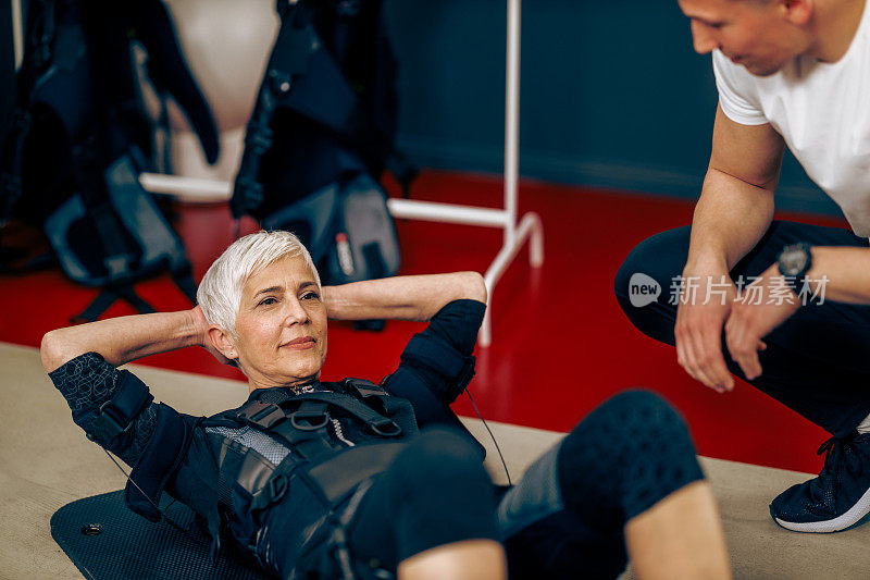 高级妇女做仰卧起坐运动期间，EMS锻炼教练在健身房