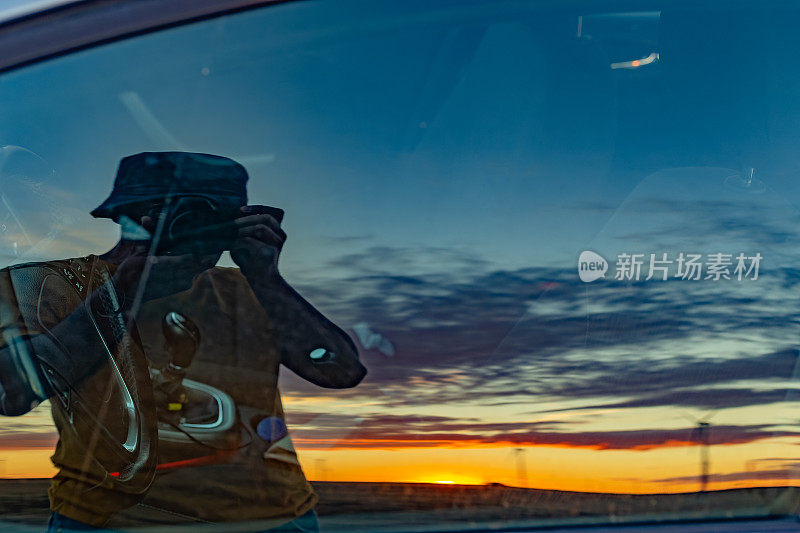 一名戴着帽子的男子在一辆汽车的驾驶座侧车窗上拍摄自己的倒影，捕捉夕阳和风力涡轮机的轮廓。
