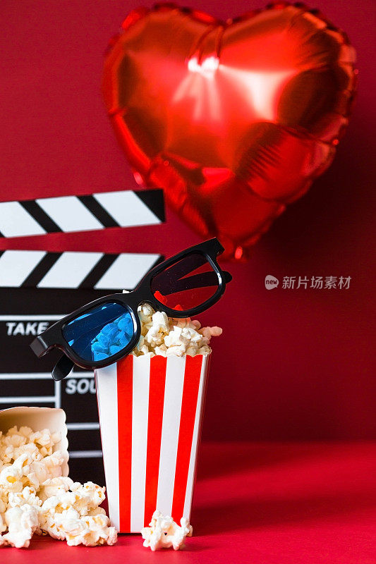 电影工业的一面旗帜。一次浪漫的电影约会。电影摄影机、3D眼镜、爆米花和红色背景上的心形铝箔气球。这部电影在情人节首映。