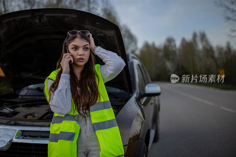 年轻的压力很大的女子站在她抛锚的汽车旁边，用手机求救