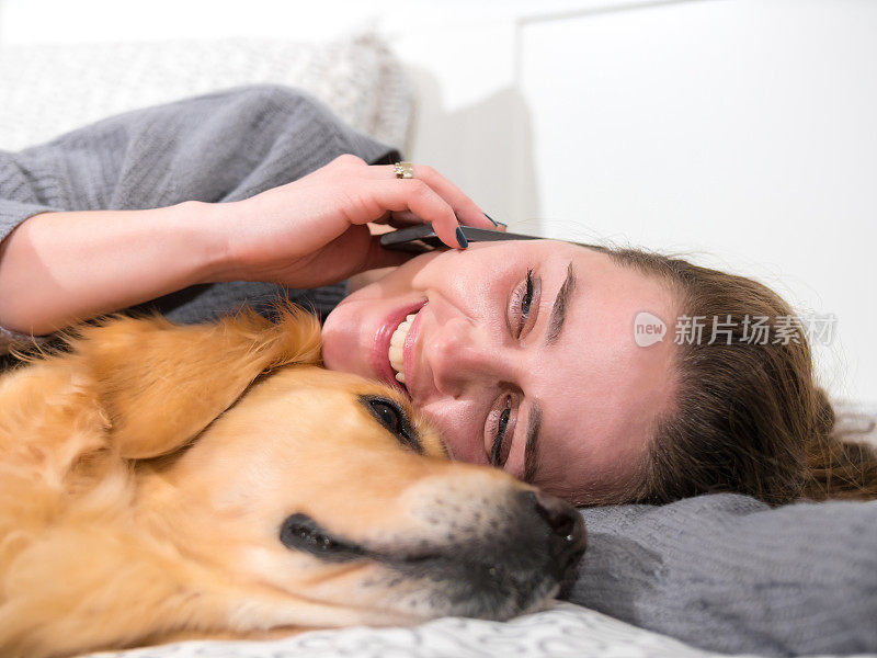 迷人的年轻女子躺在床上，旁边是她的金毛寻回犬，正在讲智能手机