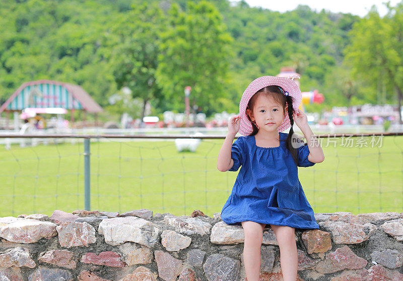 可爱的亚洲小女孩戴着草帽坐在绿色公共花园的石墙上。小女孩往外看。
