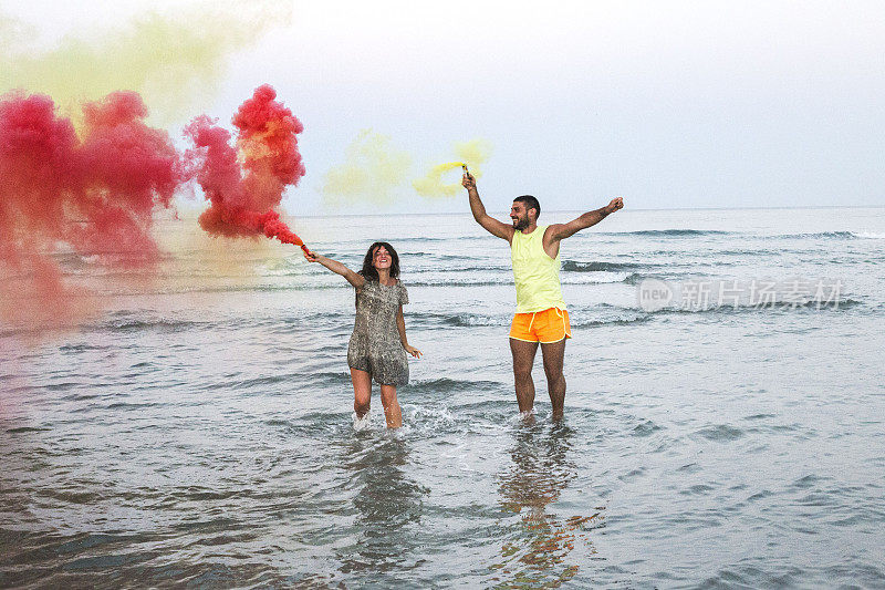 年轻的情侣站在沙滩上，手里拿着烟雾弹，浪漫的情侣用黄色和红色的烟雾弹在沙滩上