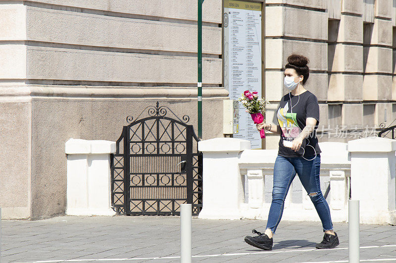 一名少女戴着口罩在一个阳光明媚的日子里走在城市街道上，手里拿着一束玫瑰
