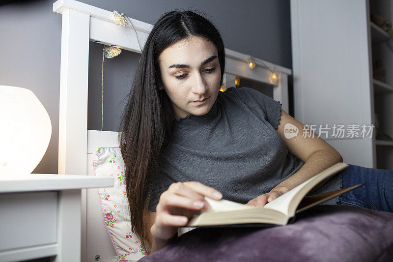 一个十几岁的女孩在她卧室的床上看书，床边的灯