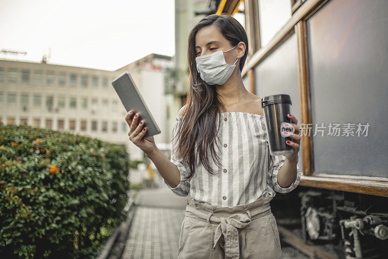 女人戴着医用口罩，用着平板电脑，喝着咖啡，在户外散步