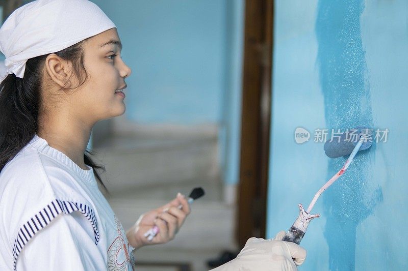 年轻女孩用油漆滚筒在墙上涂蓝色油漆。