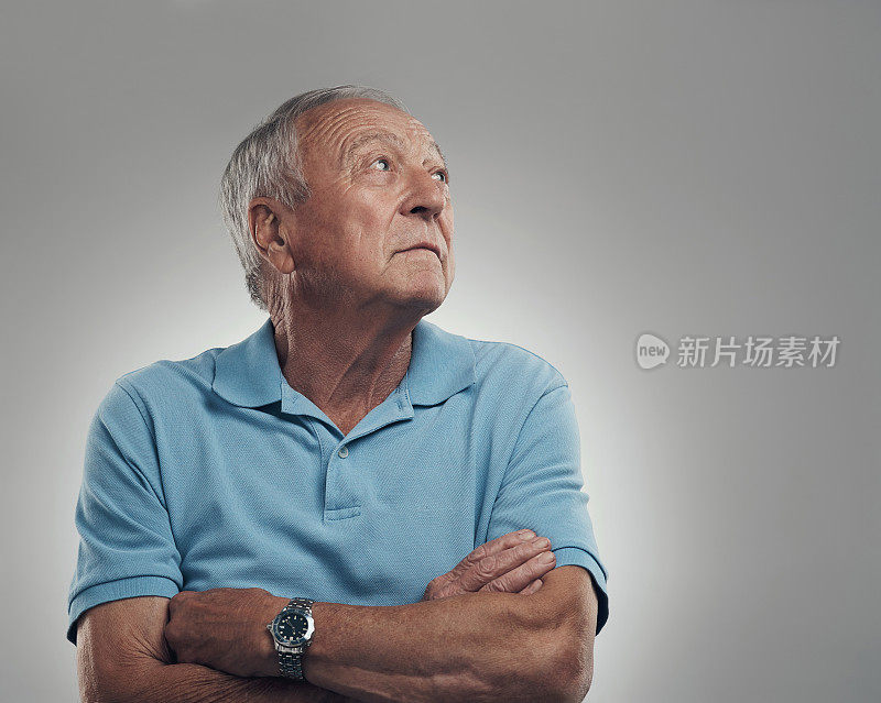 在一个灰色背景下的摄影棚里，一个年长的男人双臂交叉，望向远方