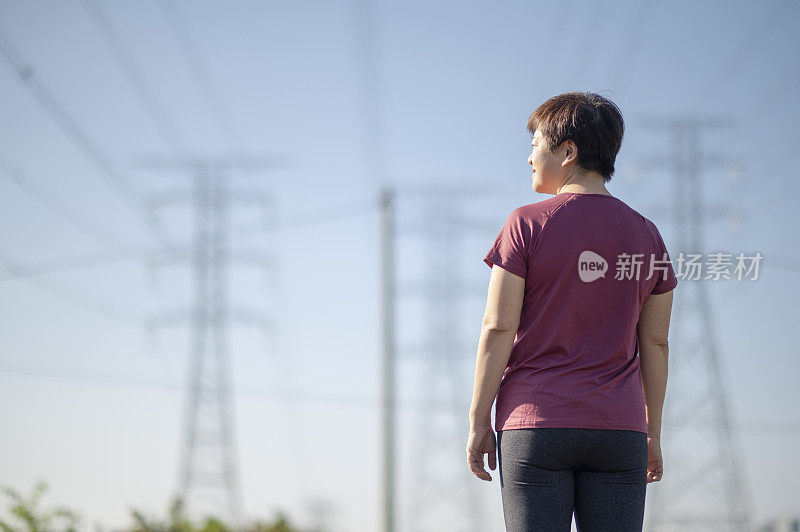 亚洲华人中年妇女与运动服装站在看在早晨与电力线蓝色的天空背景