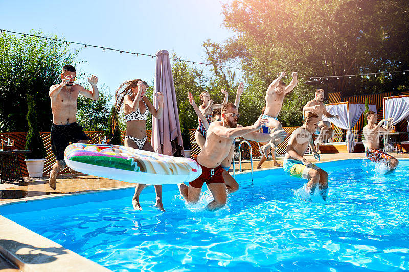快乐的朋友在豪华度假村的游泳池里跳跃和溅水花。阳光明媚的日子，年轻人穿着泳衣在私人度假别墅里聚会。辣妹跳进水里