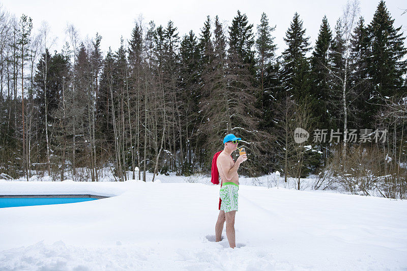 一名中年男子穿着泳裤站在那里，手里拿着一杯鸡尾酒，背景是冬天的风景。