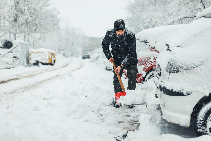 拿着雪铲的成熟男人。积雪下的汽车和街道。