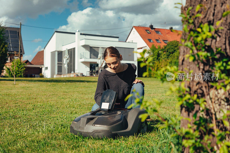 漂亮的年轻女人在家里的花园里编程机器人割草机